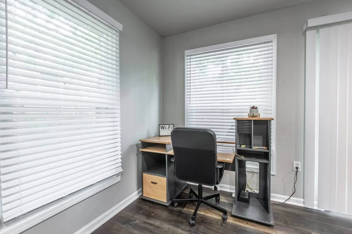This image features a dedicated workspace with a desk, office chair, blinds, and a visible Wi-Fi sign. The area is uncluttered and well-lit, clearly designed for productivity and focus, making it ideal for working, studying, or reading.
