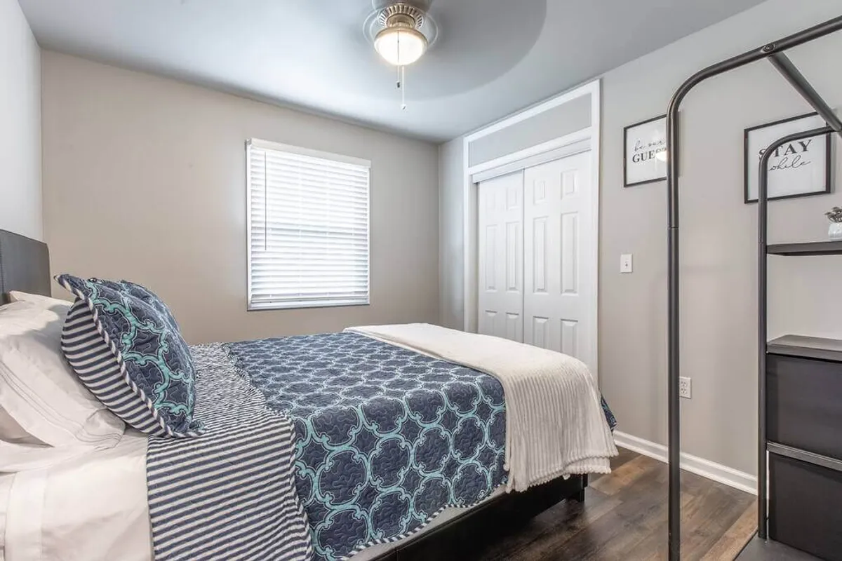A cozy and relaxing bedroom with a comfortable bed layered with decorative pillows, a nightstand with tasteful decor, and a window with blinds. The mood is intimate and inviting, ideal for reading or sleeping.