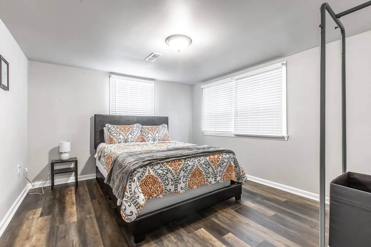 A cozy bedroom with patterned bedding, large windows letting in abundant daylight, and a nightstand with a lamp. The overall mood is calm, perfect for relaxing with a book or preparing for a productive day ahead.