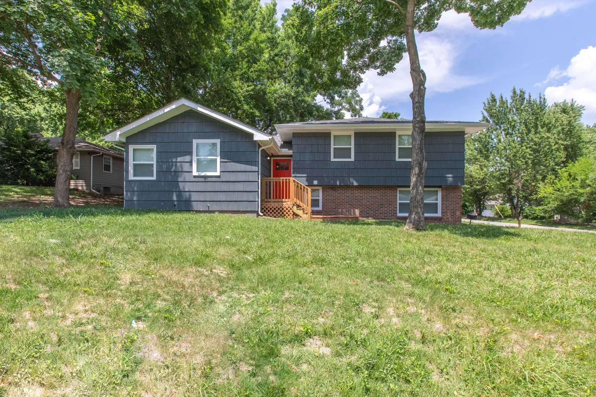 The image shows the home's inviting front porch, a green lawn shaded by mature trees, and the gray siding of the exterior. The porch offers a relaxing spot for gathering before heading out or unwinding after an event, visually connecting the home to the outdoors and neighborhood.