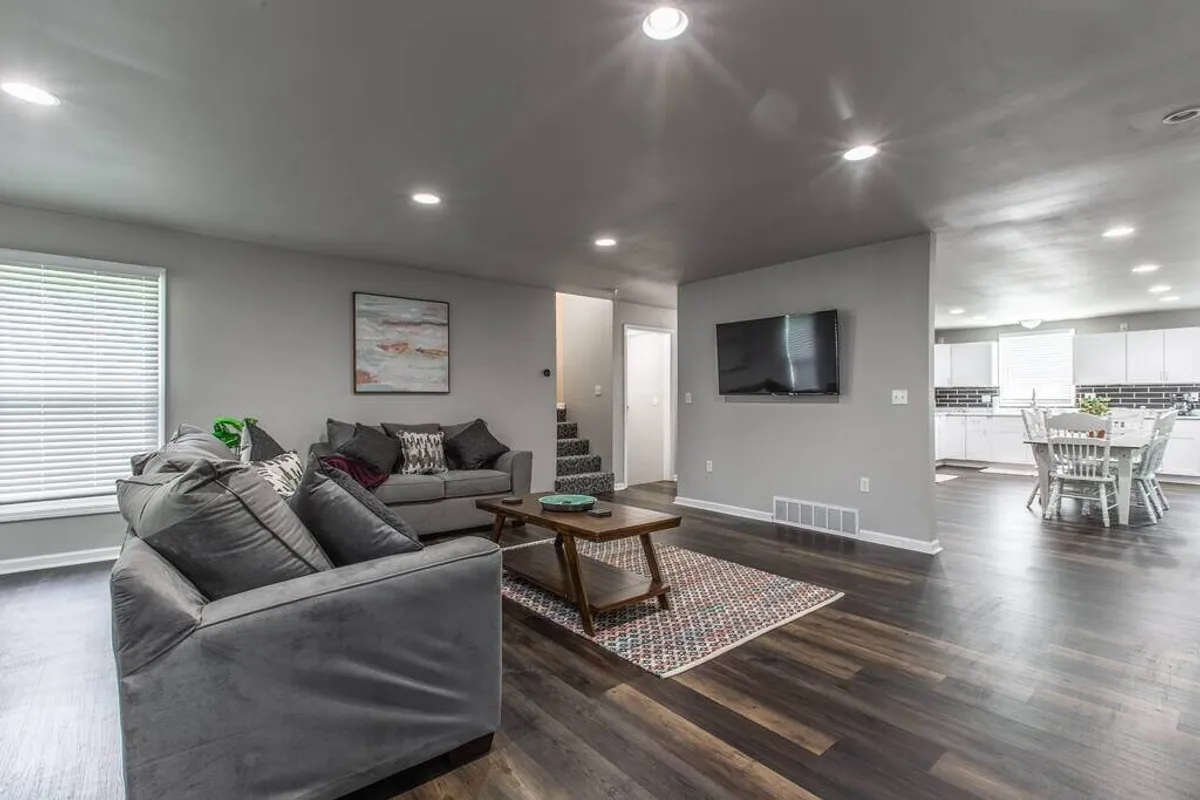 A wide view of the living room showing a comfortable sofa set, a central coffee table, a large television, and a glimpse of the open dining area and staircase. The scene is arranged for both lounging and socializing, with ample seating and a clear, inviting layout perfect for group gatherings.