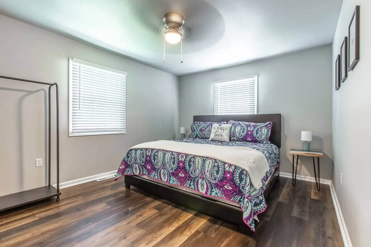 A bright, inviting bedroom featuring a bed with colorful bedding, a nightstand with a lamp, and a clothing rack. Natural light streams in from the window, creating a warm and comfortable atmosphere ideal for restful sleep or unwinding after a busy day.