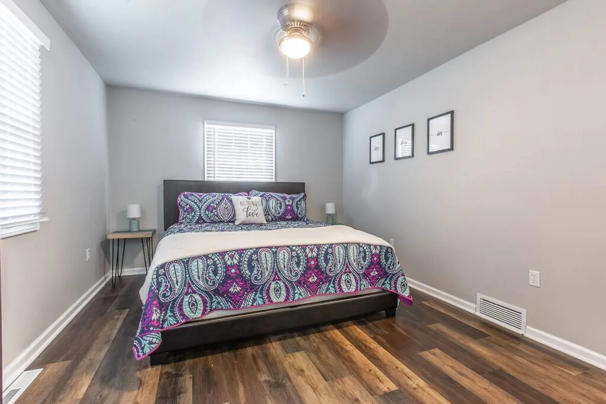 A serene bedroom featuring a bed with colorful bedding, a window with blinds softly filtering natural light, framed wall art, and nightstands with lamps on either side. The decor is calm and inviting, creating a peaceful atmosphere ideal for relaxation and restful sleep.