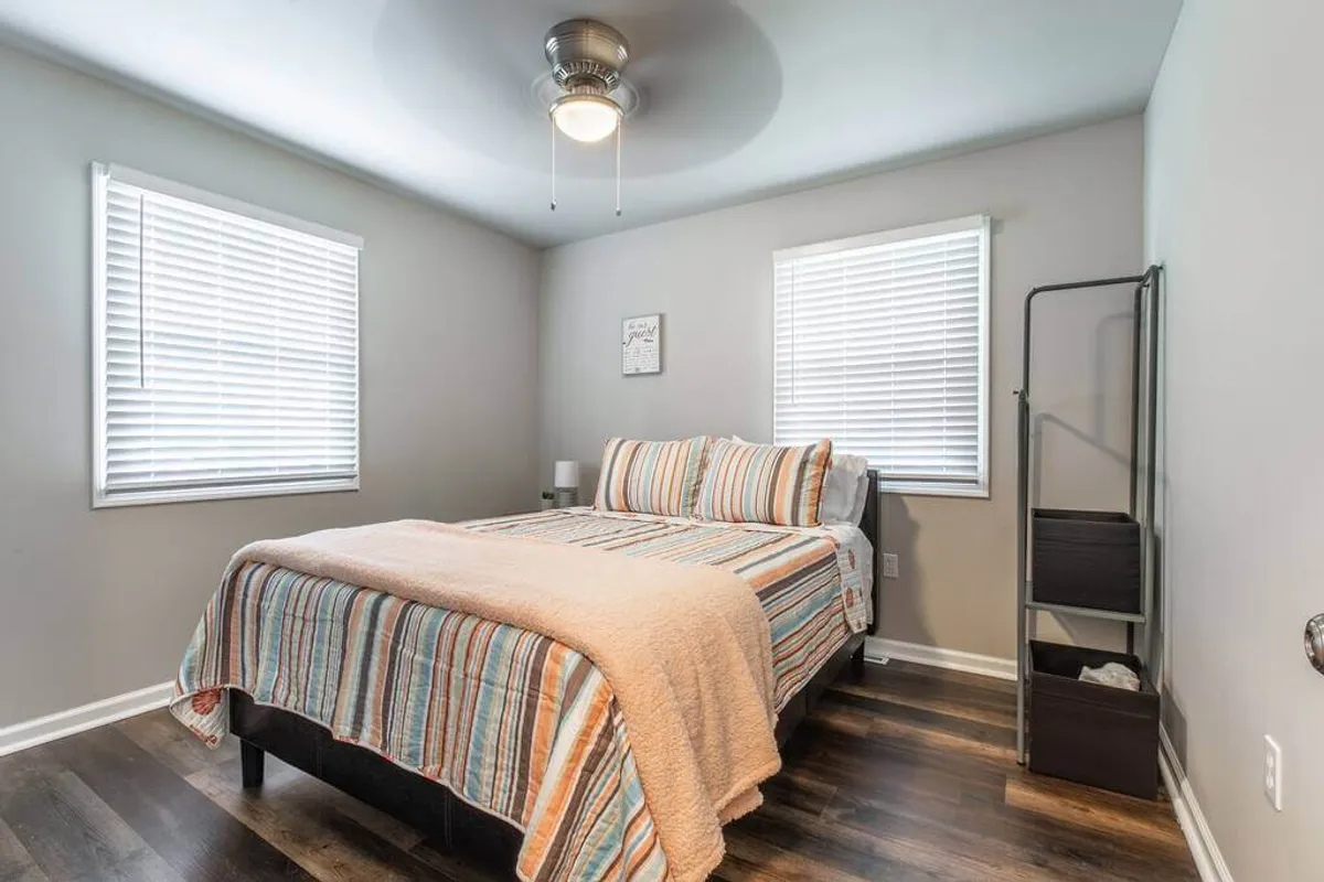A tranquil bedroom with a bed dressed in striped bedding, windows with blinds letting in gentle sunlight, a ceiling fan overhead, and a storage rack for ample storage. The muted tones and uncluttered setup highlight the room’s restful ambiance.